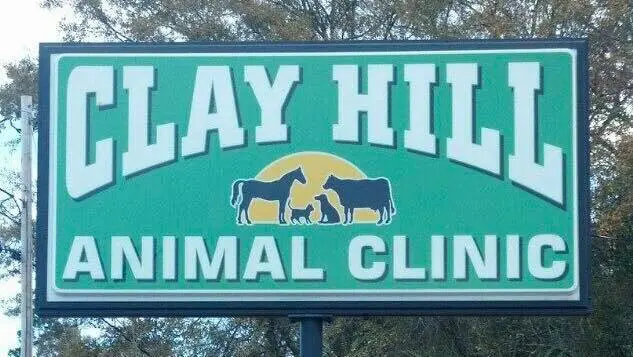 Green roadside sign reading Clay Hill Animal Clinic with a horse, cow and dogs on a yellow sun, among trees