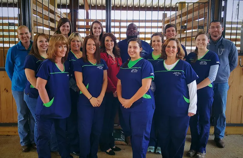 A group of smiling staff in navy scrubs with green trim standing together in front of wooden stalls