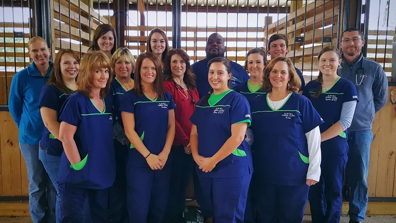 A group of smiling staff in navy scrubs with green trim standing together in front of wooden stalls